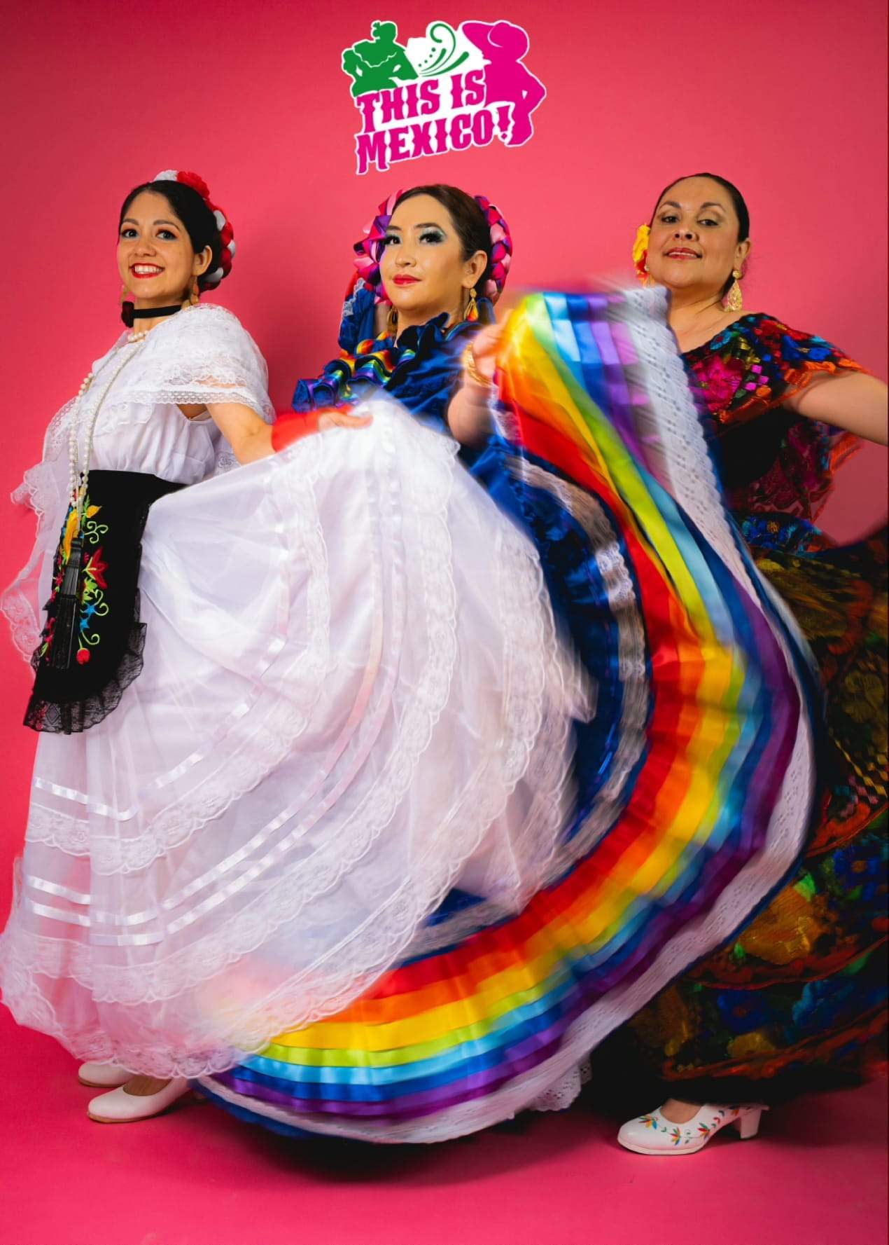 Three dancers in regional dance dress against a pink studio backdrop, skirts caught mid-turn.