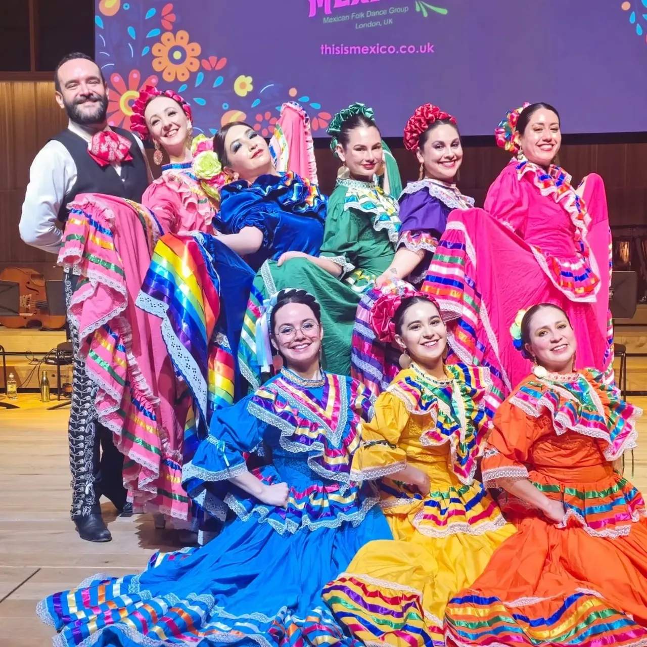 Nine dancers of This is Mexico! on stage in bright Jalisco dance dress, skirts spread, in front of the company banner.